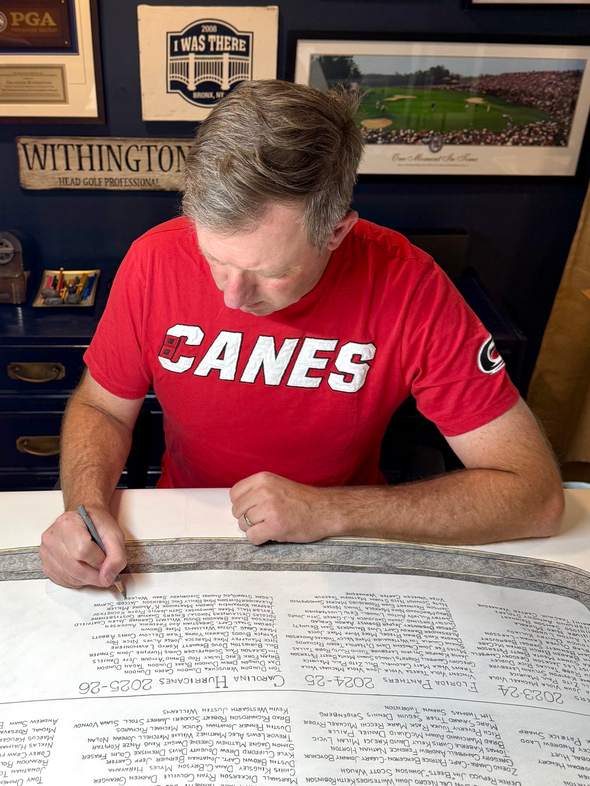 A man wearing a red t-shirt with 'CANES' printed in white across the chest sits at a desk writing on a large printed document. The background shows framed sports memorabilia on a dark blue wall.
