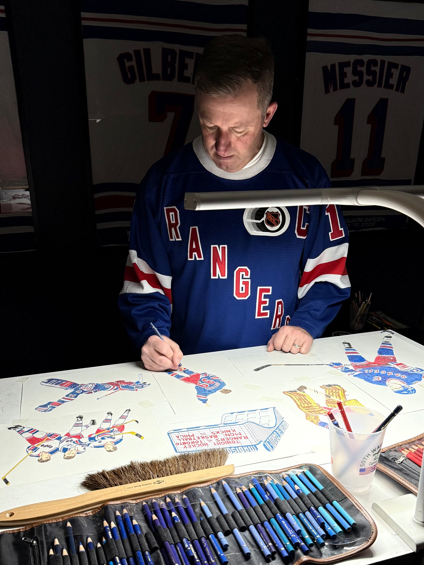 Person in a Rangers sweatshirt working on artwork with art supplies at a table.